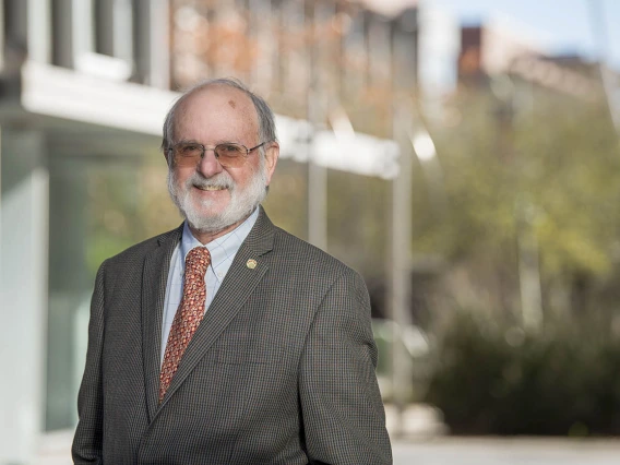 Older white man with white beard wearing a suit stands in front of a building