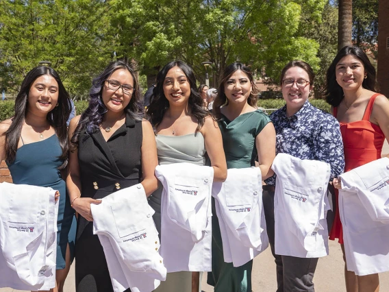 Six young women stand side-by-side outside with white coats draped over their arms. 