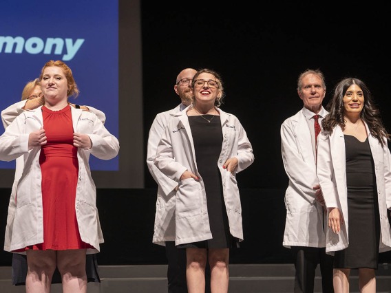 Three female Pharmacy students are presented their white coats by faculty members standing behind them on a stage. 