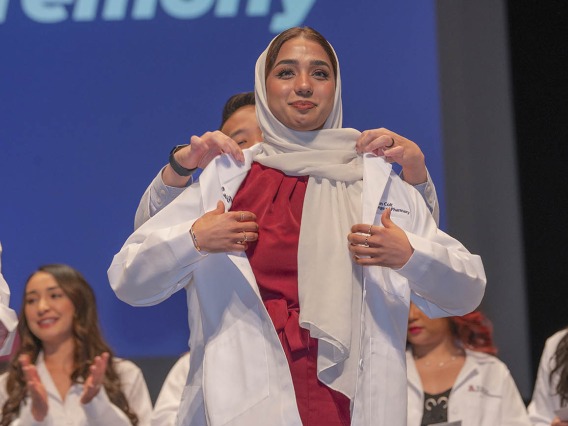 A young woman in a head scarf is presented her white coat by a faculty member standing behind her. 