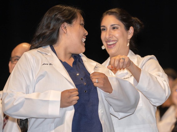 Two women wearing Pharmacy white coats smile as the younger woman puts the coat on. 