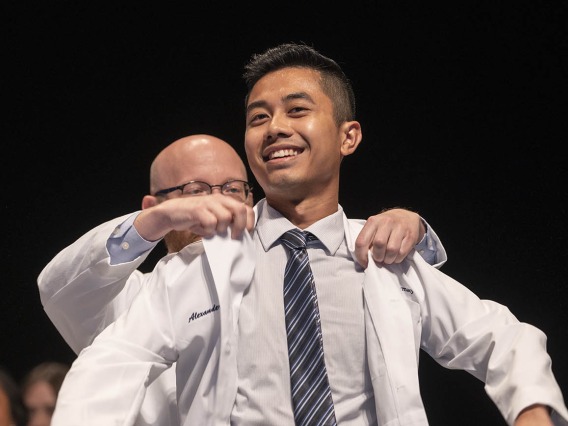 A young Asian man with a big smile is presented with his Pharmacy white coat. 