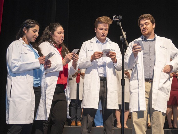 Two young women and two young men in white pharmacy coats stand in front of a microphone reading from cards they are holding. 