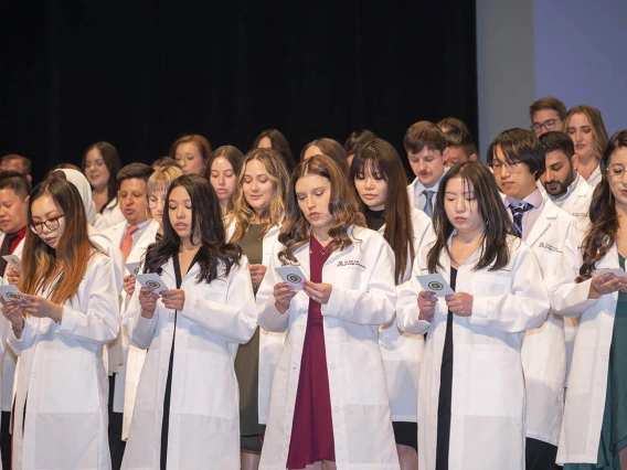 Dozens of pharmacy students wearing white coats stand on a stage reading from cards in their hands. 