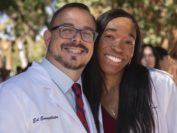 A young man and woman in pharmacy white coats smile and side-hug. 