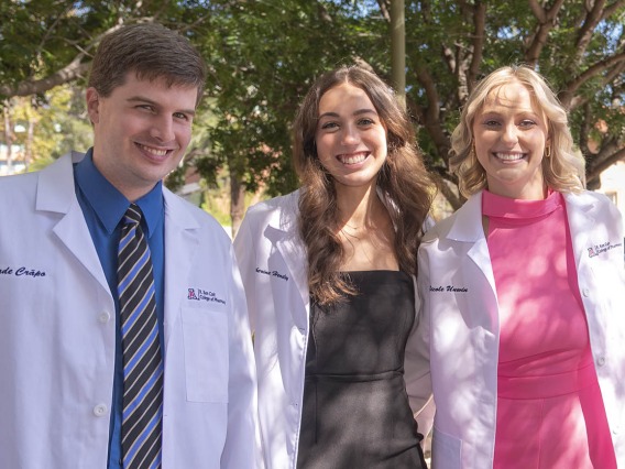Three pharmacy students wearing white coats, a male and two females, stand next to each other smiling. 