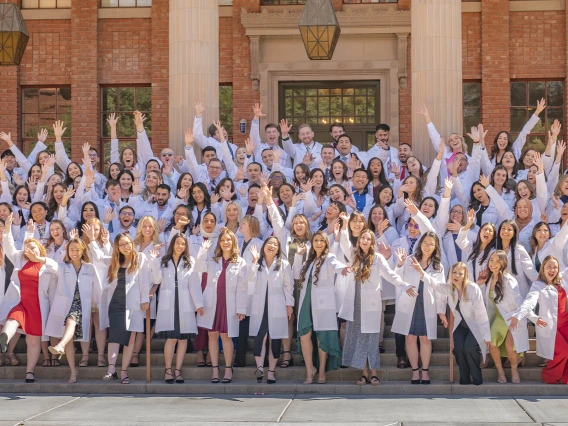 Large group of Pharmacy students wearing white coats wave their hands in celebnration.