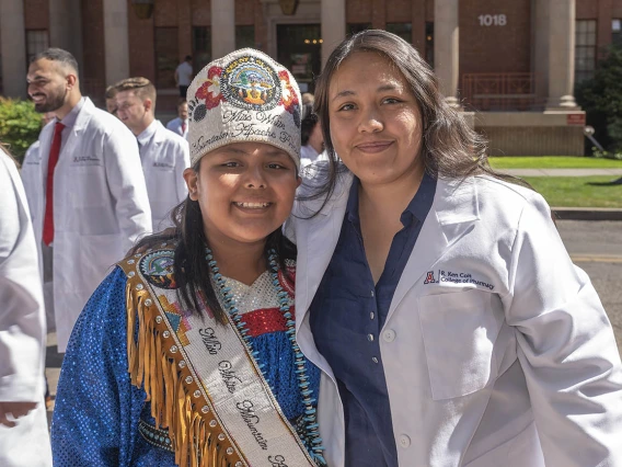 A young woman wearing traditional Native American clothing and beeded crown stands with a woman in a white pharmacy coat smiling. 