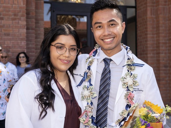 A yong woman and man wearing pharmacy white coats stand next to each other smiling. 