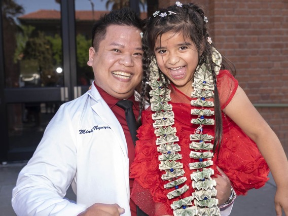 An Asian man in a pharmacy white coat holds a young girl in a red dress wearing a necklace of dollar bills. Both are smiling. 