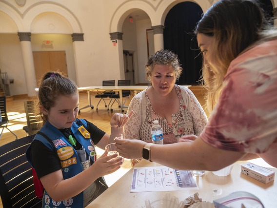 A young girl scout works with a volunteer at an activity table as her mother watches. 