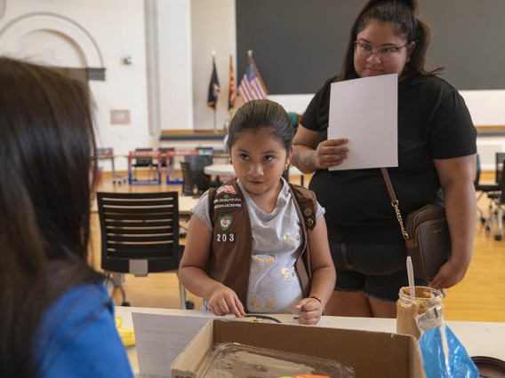 A young girl scout in uniform stands at an activity table with her mom behind her listening to a volunteer.
