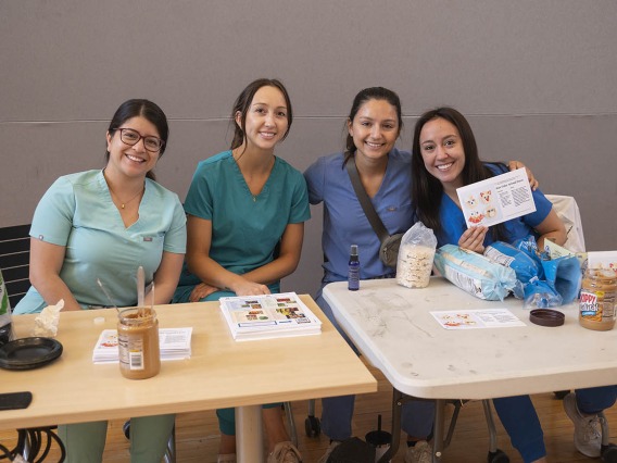 Four young women in scrubs, all medical students, sit at an activity table smiling. 