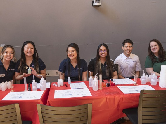 Five female and one male pharmacy students sit at an activity table smiling. 