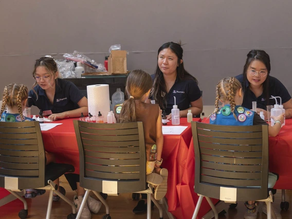 Three female pharmacy students set at a table each facing a girl scout doing an activity with them.