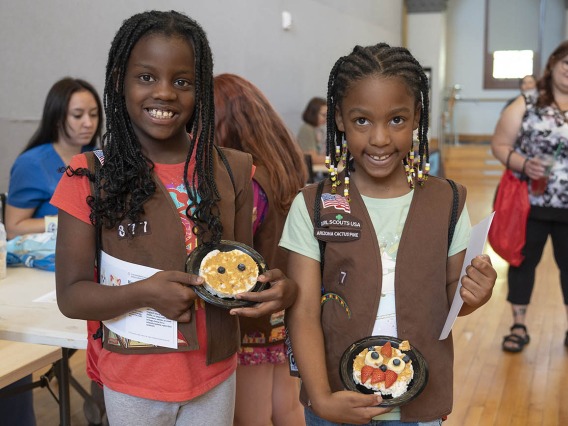 Two brown-skinned young girls in girl scout uniforms smile and hold up decorated rice cakes.