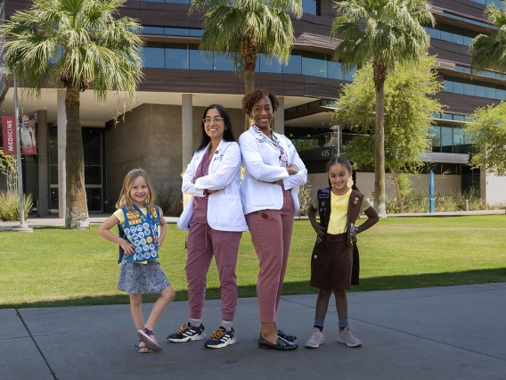 Two young girl scouts pose outside with two female medical students wearing white coats.