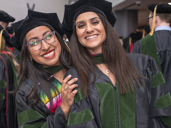 Two young women, both with long dark hair, wearing graduation regalia smile while standing closely together. 