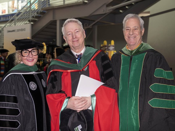 An older woman and two older men, all in graduation regalia, smile as they stand next to each other. 