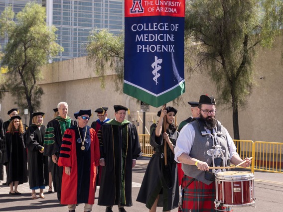 A bearded man wearing a kilt and playing a drum leads a procession of of people wearing graduation regalia down the street.