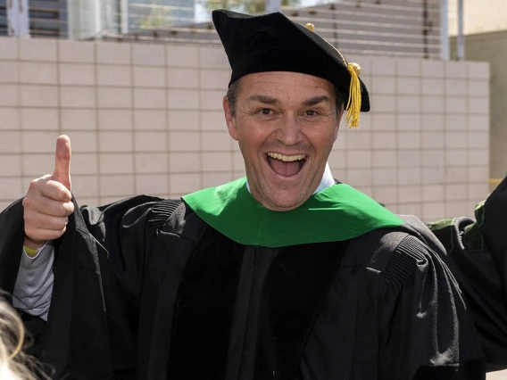 A middle-aged man wearing graduation regalia smiles and gives a thumbs-up.