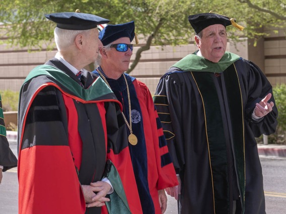 Three older men wearing graduation regalia chat while walking outside side-by-side.