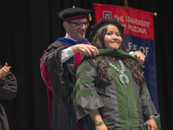 A young Native American woman dressed in graduation cap and gown has a sash placed over her shoulders by a male faculty member. 