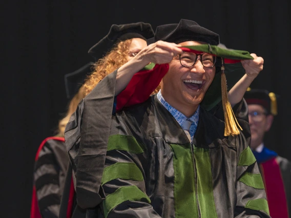 A young man with a big smile is wearing a graduation cap and gown has a sash placed over his shoulders. 