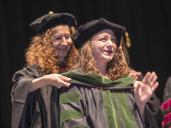 A young woman wearing graduation cap and gown waves as a sash is placed over her shoulders by a faculty member.