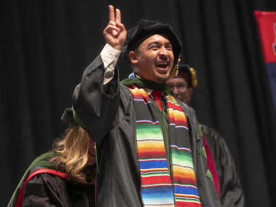 A young man wearing a graduation cap and gown smiles while holding up two fingers.