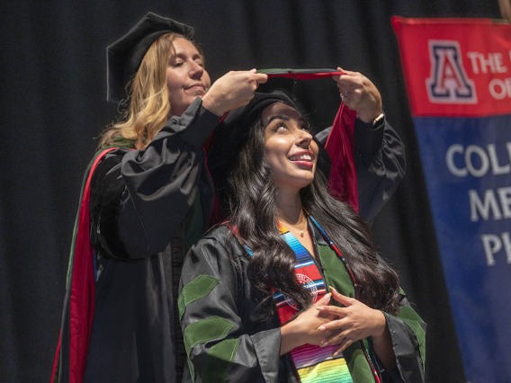 A young woman wearing a graduation cap and gown looks up and smiles as her graduation sash is placed on her shoulders by a faculty member.