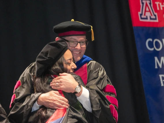 A young woman wearing graduation regalia hugs a male faculty member wearing regalia. 