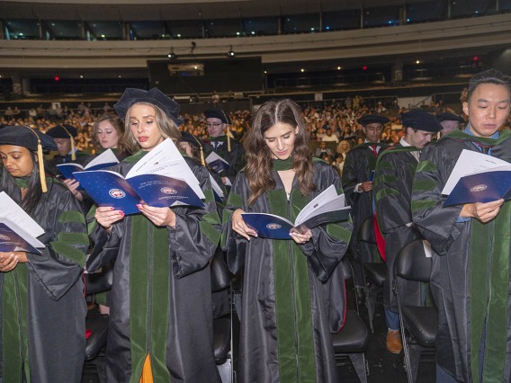 A large group of medical students wearing graduation regalia read from a pamphlet at their graduation.