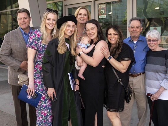 A light-skinned young blonde woman in graduation regalia stands with several members of her family, all smiling. One woman is holding a sleeping baby. 