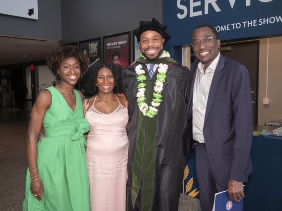 A tall dark-skinned man in graduation regalia stands with his parents and girlfriend, all smiling. 