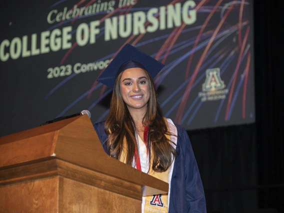 A young woman with long brown hair and dressed in cap and gown stands at a podium in Centennial Hall.