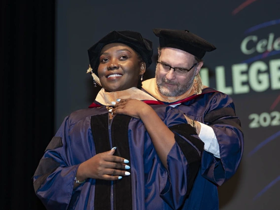 A young woman wearing graduation regalia smiles as she receives her hood by a male professor during convocation.