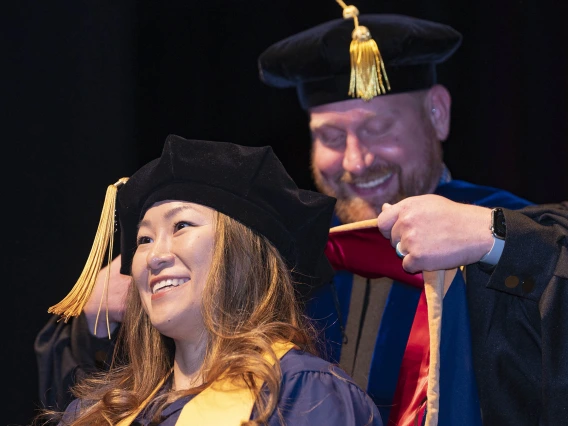 Woman with brown hair wearing graduation regalia smiles while waiting to be hooded by a male professor.