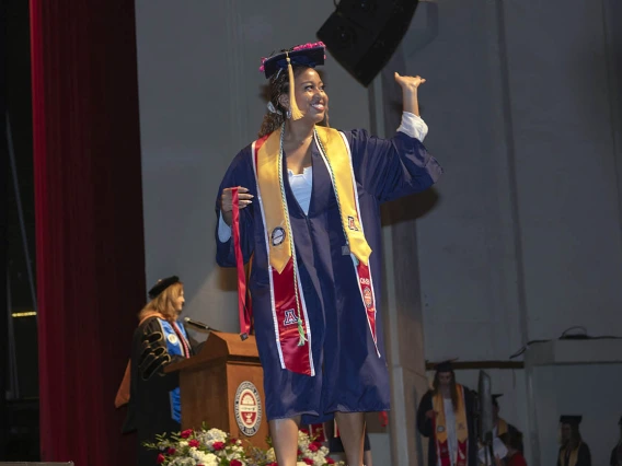 A young woman dressed in graduation regalia waves as she walks across the stage at Centennial Hall.