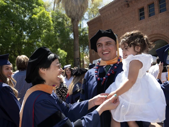 A man dressed in graduation regalia holds a toddler girl wearing a white dress. Another woman wearing graduation cap and gown reaches out to hold the toddler’s hand while smiling in celebration. 