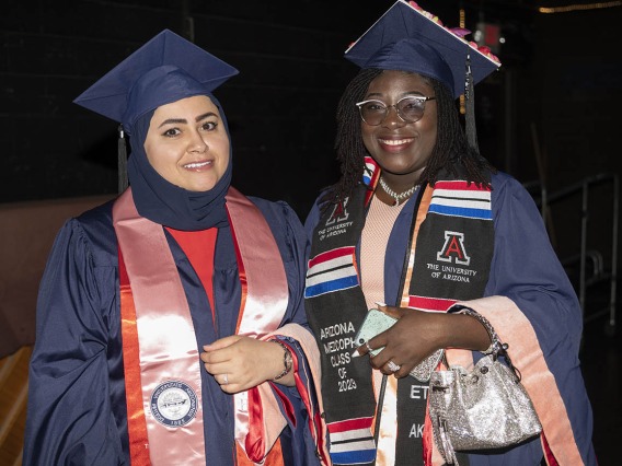 Two young women stand side-by-side wearing graduation regalia smiling. 