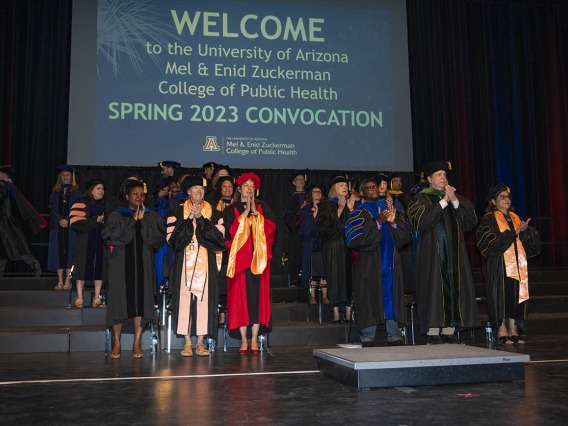 A large group of faculty members in graduation regalia stand on a stage under a large screen reading welcome to spring convocation.