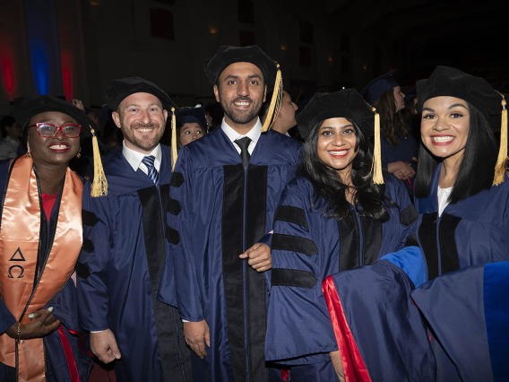 Five female and male PhD graduates in caps and gowns stand together smiling. 