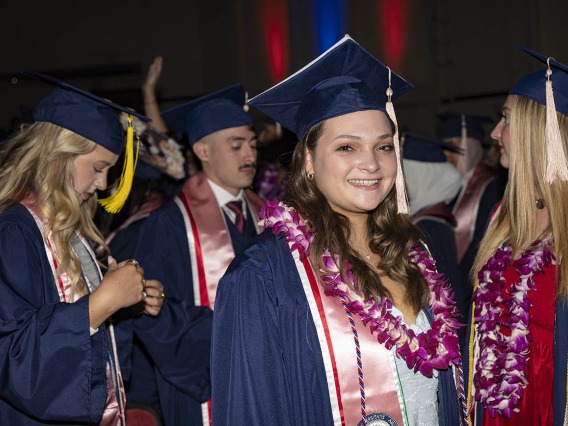A smiling young woman in a graduation cap and gown stands in front of a larger group of graduates.