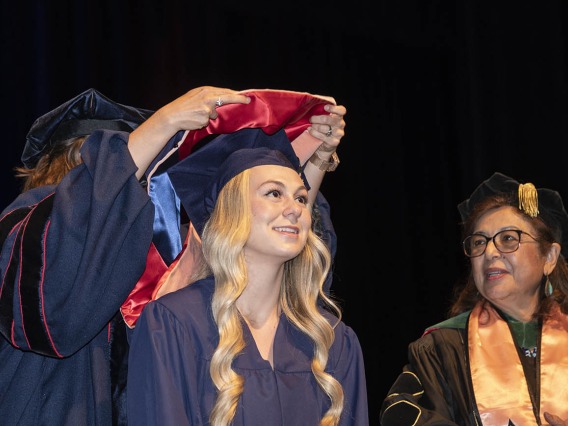 A smiling young woman with long blonde hair wearing a graduation cap and gown looks up as a professor places a sash over her shoulders. 