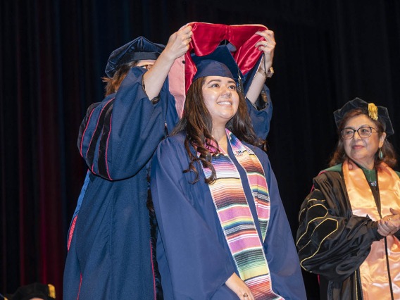 A smiling young woman in graduation cap and gown has a sash placed over her shoulders by a professor as the dean of the college looks on. 