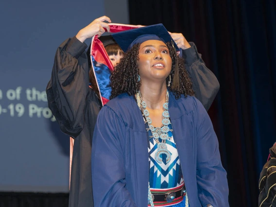 A young brown-skinned woman in graduation regalia stands smiling as a professor standing behind her places a sash over her shoulders. 