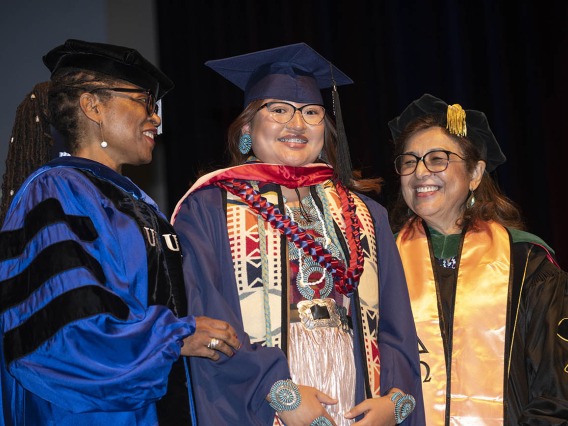A smiling young woman wearing traditional Native American clothing under her graduation cap and gown stands with two professors.
