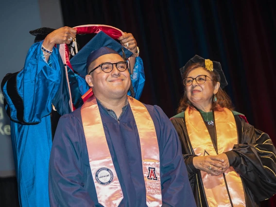 A smiling young man wearing a graduation cap and gown has a sash placed over his shoulders while the dean of the college looks on. 