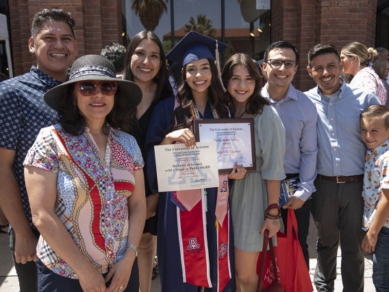 A family surrounds a young woman graduate in cap and gown holding two certificates. 